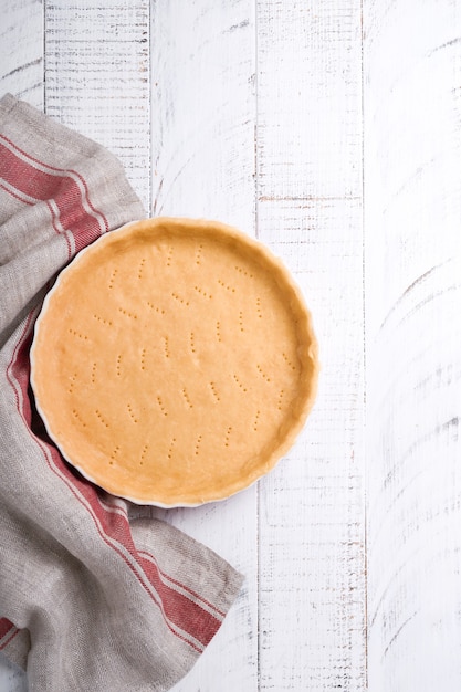 Impasto per cuocere quiche, crostata o torta in forma di cottura in ceramica pronta per la cottura su carta assorbente da cucina su fondo di legno della vecchia tavola rustica bianca. Vista dall'alto, copia dello spazio. Concetto di cottura fatta in casa per le vacanze.