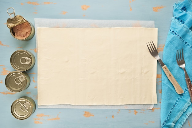 Impasto per preparare empanada ripiena di tonno su un tavolo di legno blu e scatolette di tonno