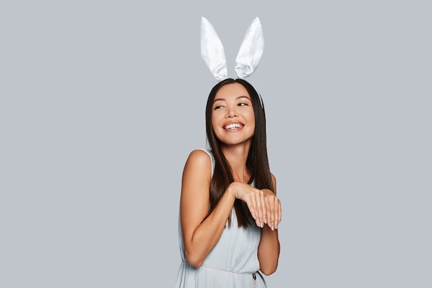 Impazzendo. Bella giovane donna asiatica con orecchie da coniglio che sorride mentre sta in piedi su uno sfondo grigio