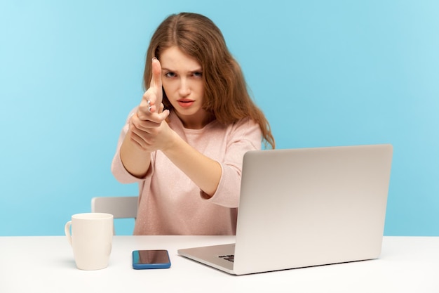 Impiegata arrabbiata seduta sul posto di lavoro con laptop che minaccia e punta la pistola del dito verso la telecamera con espressione aggressiva mirando al bersaglio tiro in studio indoor isolato su sfondo blu