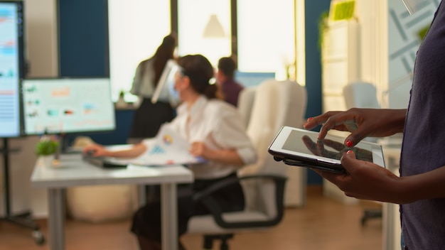 Impiegato africano che utilizza tablet in piedi nella stanza dell'ufficio e team di analisti finanziari che lavorano in background. Colleghi multietnici che rispettano la distanza sociale in un'azienda durante la pandemia globale