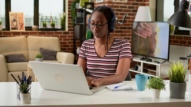 Impiegato afroamericano che lavora in remoto e ascolta musica con le cuffie. Donna d'affari che lavora a distanza sul laptop, utilizzando la connessione di rete, indossando l'auricolare alla scrivania di casa.