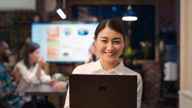 Impiegato asiatico che lavora al computer portatile, che guarda l'obbiettivo, sorridente, rallentatore. Giovane donna d'affari in possesso di computer vicino ritratto, presentazione riunione d'affari in background. Colpo a mano libera.