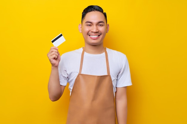 Impiegato barista sorridente di successo giovane uomo asiatico 20s che indossa grembiule marrone che lavora nella caffetteria in possesso di carta di credito guardando la fotocamera su sfondo giallo Concetto di avvio di piccole imprese