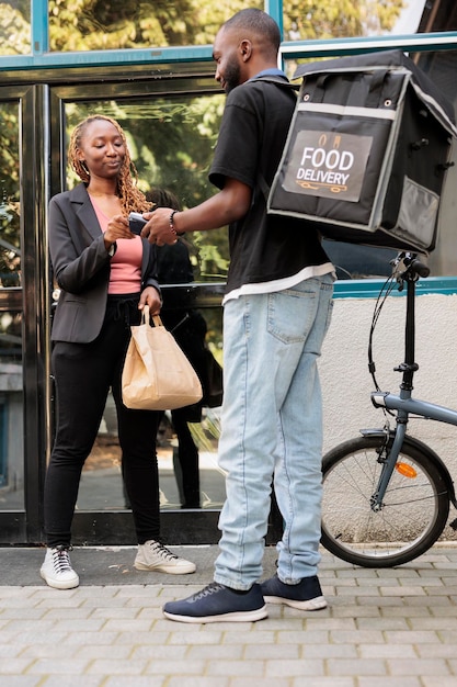 Impiegato che paga la consegna del cibo con carta di credito, terminale pos pagamento senza contatto per l'ordine. Corriere afroamericano con zaino che consegna il pasto al ristorante al cliente all'aperto