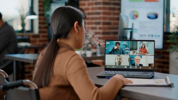 Impiegato dell'azienda che utilizza la videoconferenza online sul laptop per parlare con gli uomini d'affari in ufficio. Lavoratrice in sedia a rotelle che si riunisce in teleconferenza remota per la comunicazione a distanza.