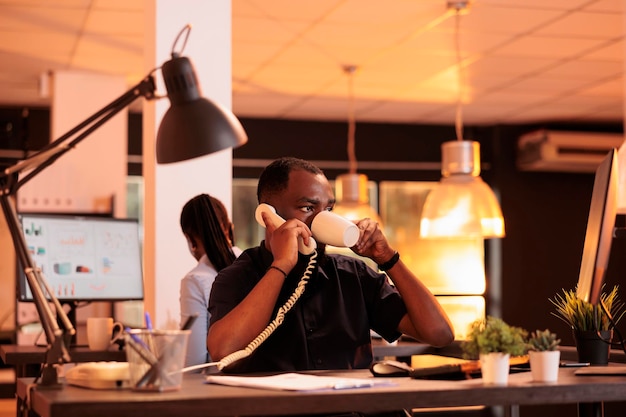 Impiegato di avvio che utilizza il telefono fisso per avere una conversazione remota durante il tramonto. Chiacchierando sul telefono dell'ufficio con i colleghi per lavorare su rapporti di lavoro e stipulare contratti di scartoffie.