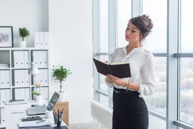 Impiegato femminile che annota note e programma giornaliero nel suo taccuino, in piedi sul posto di lavoro, ritratto vista frontale.