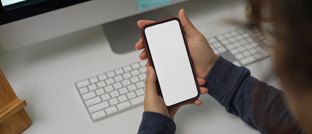 Impiegato femminile che si rilassa con lo smartphone dello schermo in bianco mentre lavorando con il computer sulla scrivania