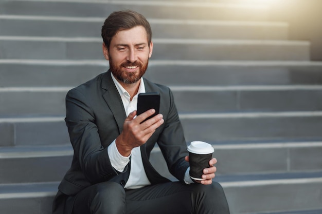 Impiegato sorridente in tuta seduto sulle scale con caffè durante la pausa e usando il telefono