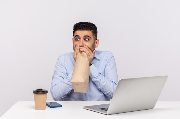 Impiegato spaventato seduto sul posto di lavoro dell'ufficio con il laptop, espirando e inalando nella confezione, usando il sacchetto di carta per migliorare il benessere, superando la paura dello stress sul lavoro. tiro in studio indoor isolato