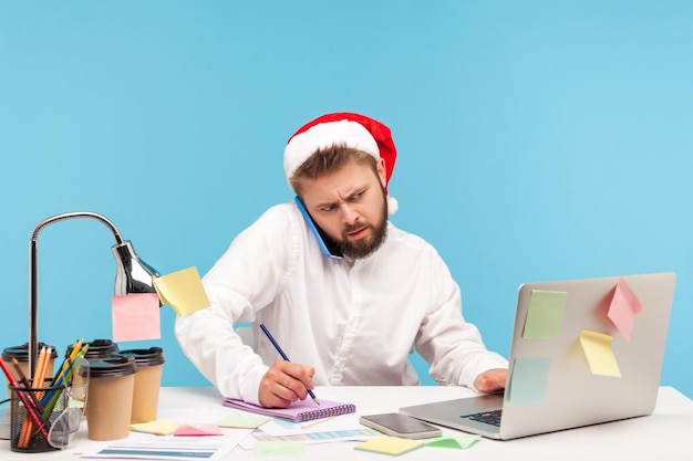 Impiegato uomo concentrato seduto sul posto di lavoro con il laptop in cappello di Babbo Natale, parlando al telefono e prendendo appunti sul blocco note, lavorando nei giorni festivi. Studio indoor girato isolato su sfondo blu