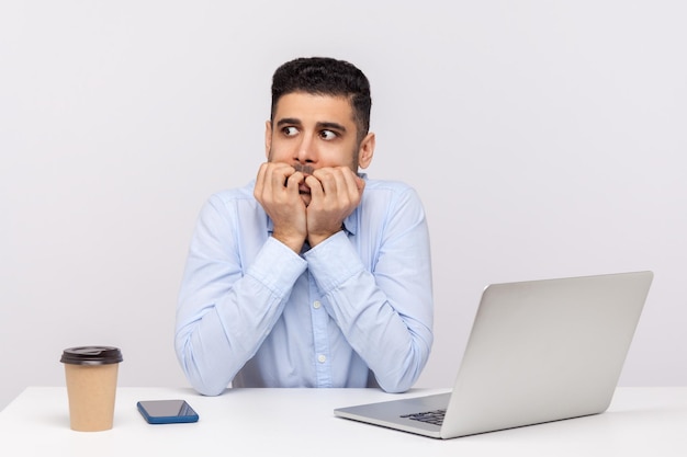 Impiegato uomo nervoso spaventato seduto sul posto di lavoro in ufficio con laptop e caffè, mangiarsi le unghie, in preda al panico terrorizzato dalla scadenza dei problemi, disturbo d'ansia, lavoro stressante. tiro in studio indoor isolato