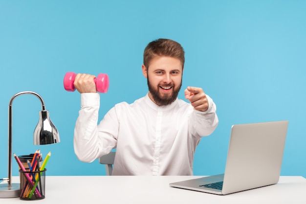 Impiegato uomo sano positivo che tiene il manubrio e punta il dito su di te, facendo ginnastica sul posto di lavoro, flettendo i muscoli durante lo stile di vita sedentario. Studio indoor girato isolato su sfondo blu