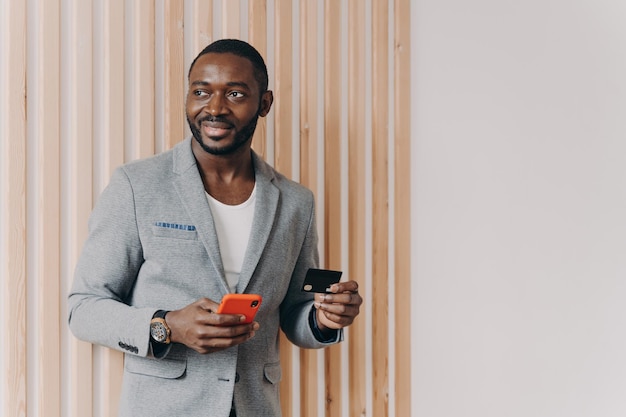Imprenditore afroamericano allegro in giacca elegante con carta di credito e smartphone in mano