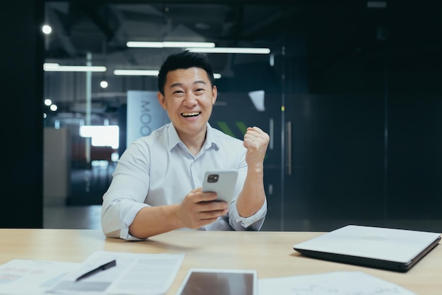Imprenditore asiatico di successo con il telefono che guarda l'obbiettivo e sorride celebrando l'uomo della vittoria a