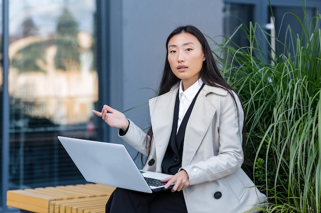 Imprenditore di successo donna asiatica che lavora su laptop all'aperto seduto vicino al centro ufficio e guardando la fotocamera