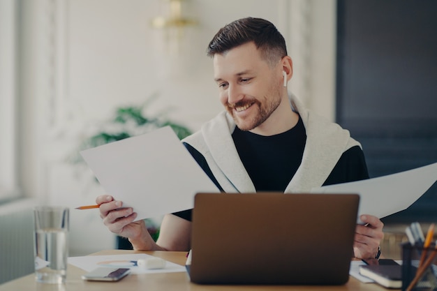 Imprenditore di successo in auricolari wireless, analizzando documenti e guardando soddisfatto dei risultati aziendali mentre si lavora da casa. Impiegato maschio seduto al suo posto di lavoro e sentirsi felice