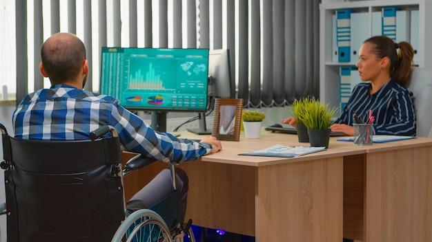 Imprenditore disabile seduto in sedia a rotelle immobilizzato analizzando le statistiche dell'economia finanziaria sul computer in ufficio che discute con il collega. Uomo d'affari disabile che utilizza la tecnologia moderna
