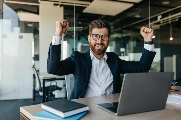 Imprenditore emotivo gesticolando sì e agitando i pugni seduto sul posto di lavoro in ufficio e sorridendo alla telecamera