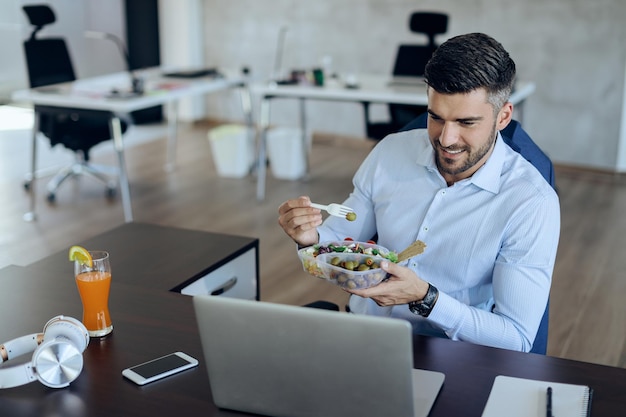 Imprenditore felice che mangia cibo sano mentre utilizza il computer in ufficio