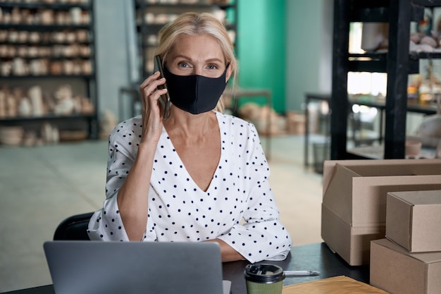 Imprenditore femminile che indossa una maschera protettiva che parla al telefono e lavora al computer portatile