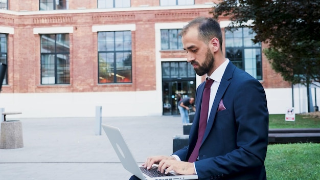 Imprenditore in abito diplomatico che tiene in mano un computer portatile che lavora alla presentazione aziendale digitando la presentazione della gestione. Direttore esecutivo seduto sulla panchina di fronte all'ufficio di costruzione di avvio