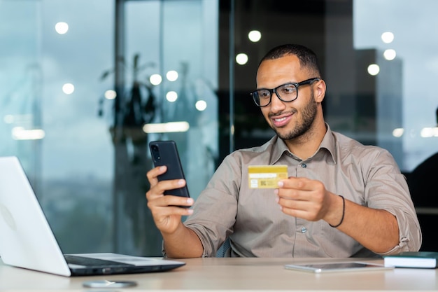 Imprenditore ispanico di successo all'interno dell'uomo dell'ufficio in camicia sorridente e felice in possesso di credito bancario