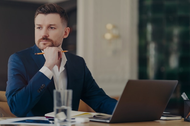 Imprenditore maschio impegnato in abito blu scuro seduto alla sua scrivania in un ufficio elegante e pensando a nuove soluzioni alle sfide aziendali, tenendo in mano la matita pur essendo serio e concentrato