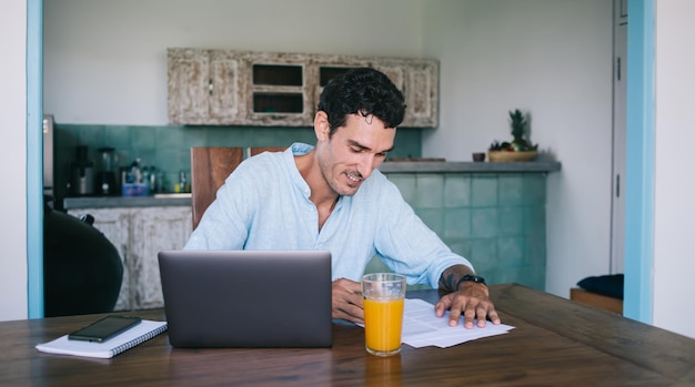 Imprenditore maschio sorridente entusiasta in camicia leggera seduto al tavolo e bere succo mentre analizza le vendite completate nei documenti in una cucina accogliente