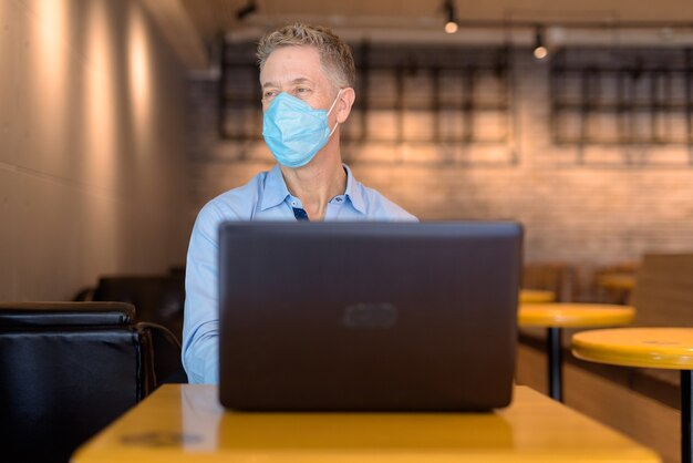 Imprenditore maturo con maschera pensando durante l'utilizzo di laptop presso la caffetteria