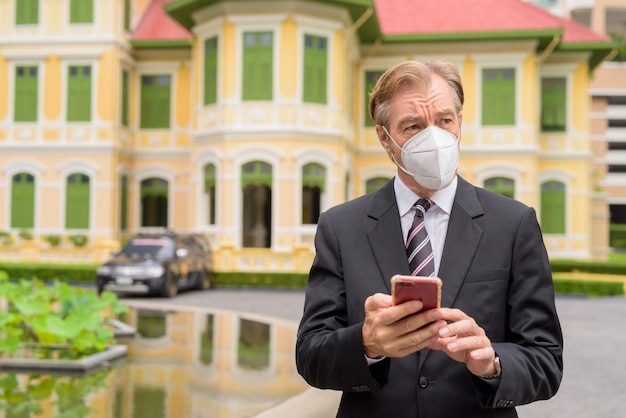 Imprenditore maturo con maschera pensando mentre si utilizza il telefono in città all'aperto