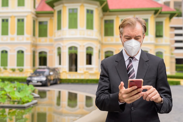 Imprenditore maturo con maschera utilizzando il telefono in città all'aperto