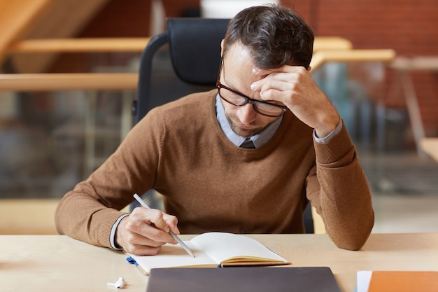 Imprenditore maturo in occhiali seduto al tavolo e prendere appunti nel blocco note ha pianificato la sua settimana lavorativa in ufficio