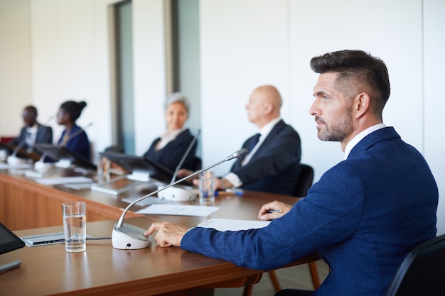 Imprenditore maturo seduto al tavolo utilizzando il microfono per parlare un discorso per uomini d'affari durante la conferenza