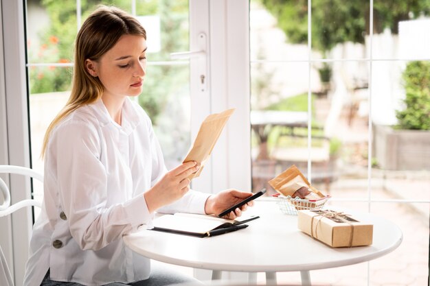 Imprenditore online Le donne caucasiche indossano una camicia bianca, guardano lo smartphone, controllano l'ordine, molte scatole di pacchi con pacchi e carrello sul tavolo nella caffetteria vicino alla finestra. concetto di vendita