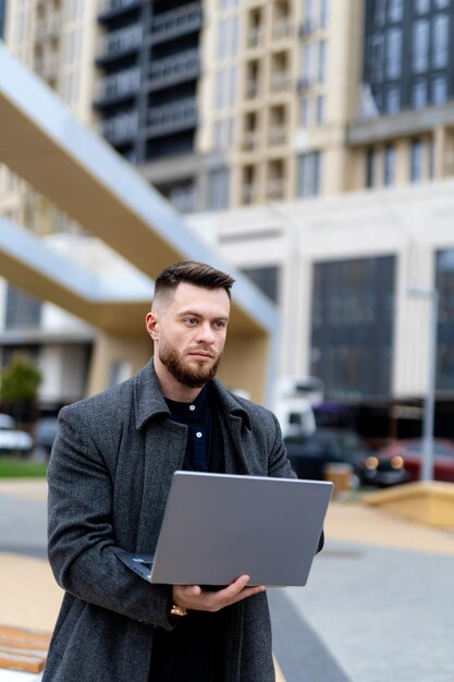 Imprenditore persistente in cappotto grigio casual che lavora su laptop Strada ampia e moderna sullo sfondo