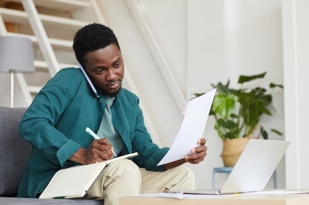 Imprenditore serio africano seduto sul divano davanti al computer portatile e facendo il lavoro di ufficio che lavora online a casa