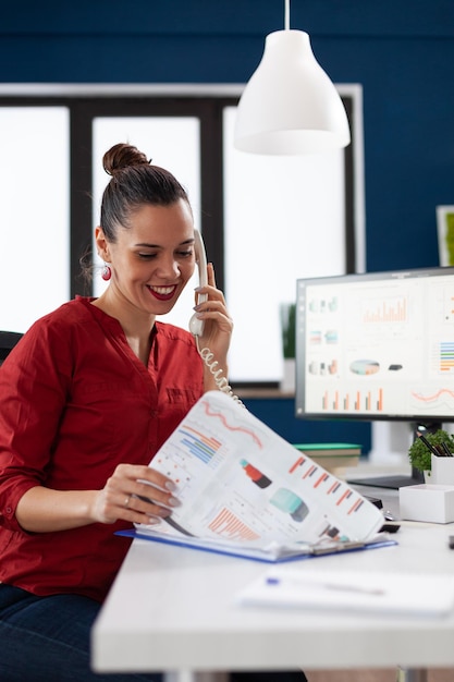 Imprenditore sorridente che parla al telefono spiegando i grafici. Manager di successo soddisfatto dei risultati aziendali nell'ufficio di avvio. Impiegato in camicia rossa che lavora alla scrivania con appunti e computer.