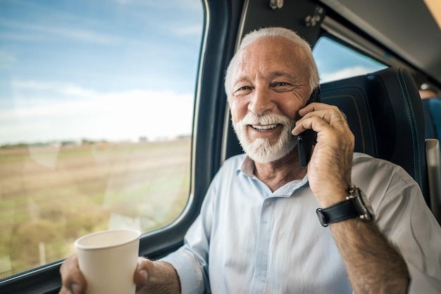 Imprenditore sorridente dell'uomo d'affari che parla sullo Smart Phone mentre beve caffè sul treno intercity