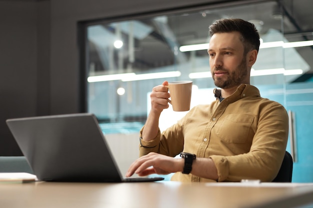 Imprenditore utilizzando laptop e bere caffè lavorando seduto in ufficio