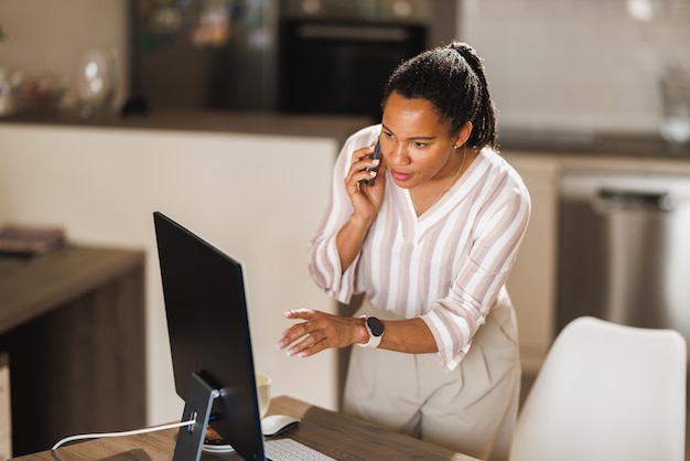 Imprenditrice africana che utilizza un telefono cellulare mentre lavora al computer dal suo ufficio a casa.