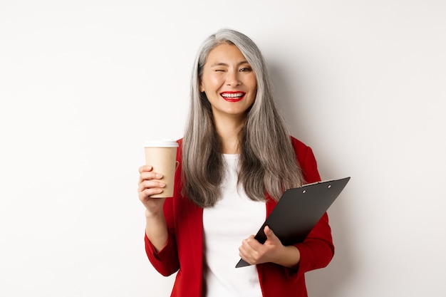 Imprenditrice asiatica di successo che beve caffè e strizza l'occhio allegramente alla telecamera, in piedi con gli appunti, indossa un blazer rosso, sfondo bianco