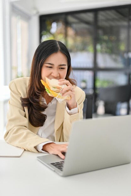 Imprenditrice asiatica impegnata e laboriosa che mangia un panino mentre lavora al suo progetto