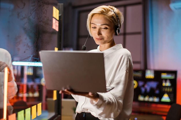 Imprenditrice che lavora in ufficio vicino alla parete di vetro utilizzando cuffie e laptop