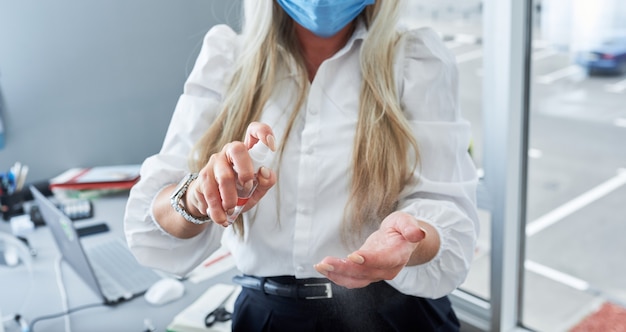 Imprenditrice con maschera e guanti utilizzando disinfettante per le mani in ufficio. Protezione dal coronavirus.