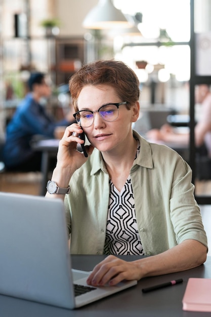 Imprenditrice con una conversazione sul cellulare mentre si lavora online sul laptop seduto al tavolo in ufficio