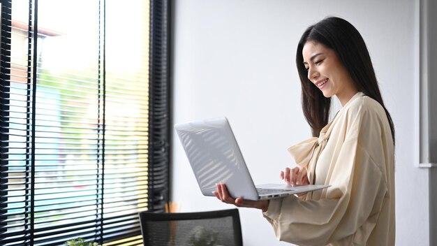 Imprenditrice di successo in piedi vicino alla finestra dell'ufficio e usando il suo laptop