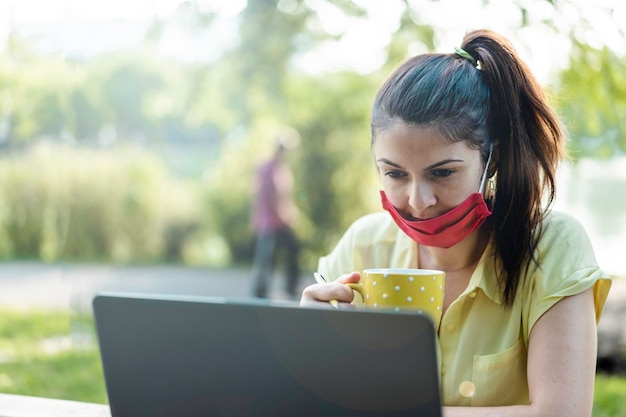 Imprenditrice femminile che indossa una maschera protettiva aperta utilizzando il laptop mentre beve caffè all'aperto Donna che lavora in uno spazio di lavoro alternativo al parco che lavora a distanza nella natura dopo la pandemia di covid19