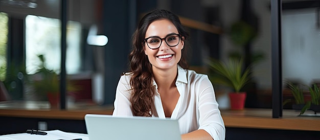 Imprenditrice femminile che lavora alla scrivania in ufficio sorridendo alla telecamera con spazio vuoto sullo schermo del laptop per la copia